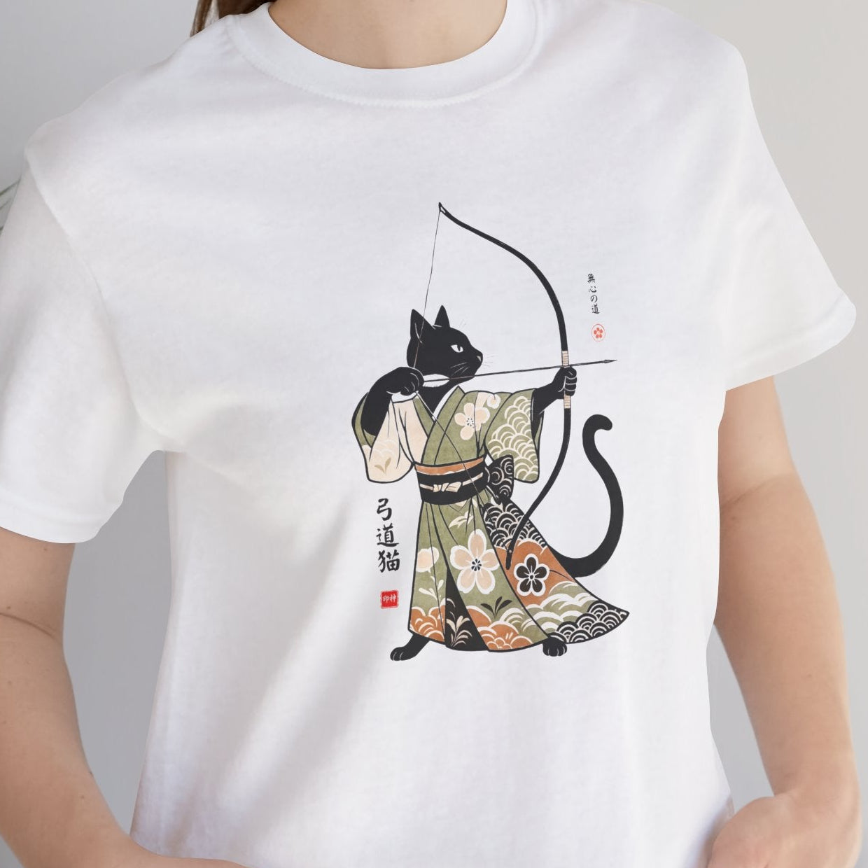 Person wearing a white t-shirt with a black cat in a kimono design, standing indoors next to a plant.